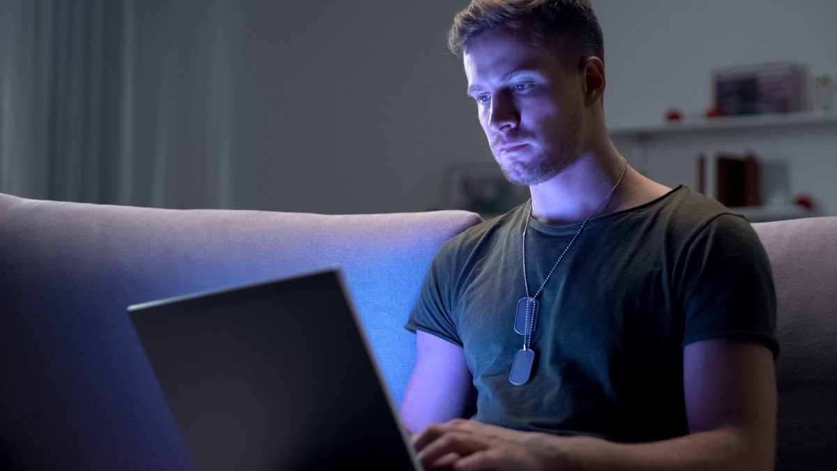Young adult male with army dog tags working on laptop in dark room