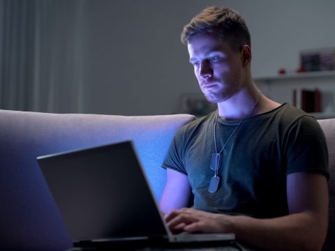 Young adult male with army dog tags working on laptop in dark room